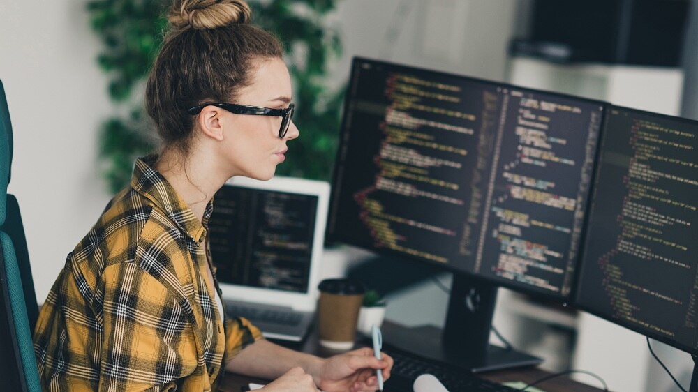 Eine professionelle aussehende junge Frau in legerer Kleidung, konzentriert auf das Programmieren an zwei Bildschirmen in einer Remote-Arbeitsumgebung.